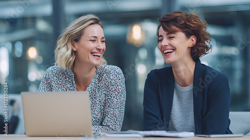 Two smiling women at a table one with a laptop in a modern office setting