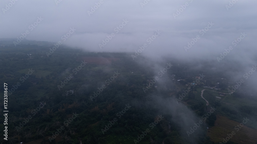 Fototapeta premium Aerial of Village in rain cloud cover tropical green mountain. Rainy season. Misty cover green forest. beautiful green village