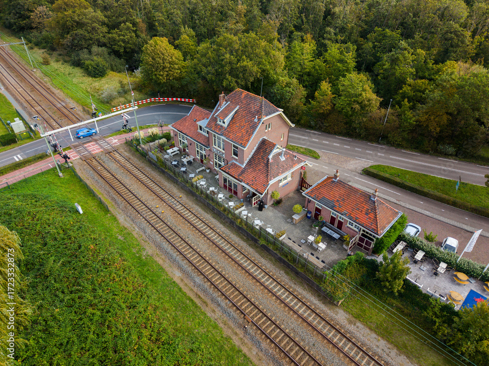 Fototapeta premium Aerial view of a red roofed cafe beside railway tracks and a road crossing with safety barriers, set in a green semi rural landscape of trees and vegetation.