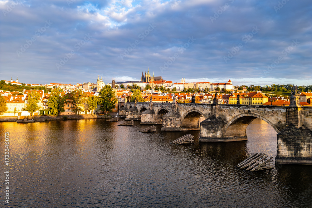 Fototapeta premium Early morning light casts a glow on Prague Castle and Charles Bridge, reflecting beauty over the Vltava River. The old town awakens as clouds drift above the historic skyline.