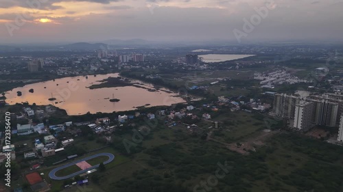 An aerial shot captures a dramatic sunset over a serene lake, contrasting with the signs of urban expansion. The footage showcases distant high-rise buildings and new land plots, Sport track Highriser