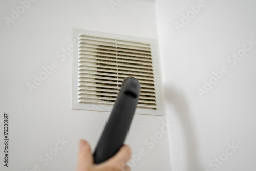 A hand holds a vacuum cleaner attachment directed towards a vent cover while performing cleaning tasks to improve indoor air quality in a residential space.