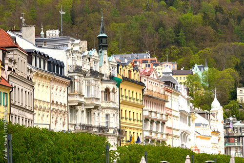 This is a vibrant view of Karlovy Vary, showing a row of colorful, ornate buildings nestled against a green, forested hillside. The architecture is a mix of styles