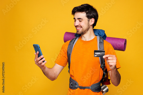 Young man carry backpack with stuff mat use mobile cell phone credit card isolated on plain yellow background. Tourist lead active lifestyle walk on spare time. Hiking trek rest travel trip concept.