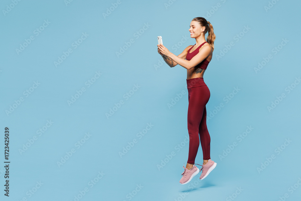 Obraz premium Full body young fitness trainer instructor sporty woman wear red sportswear train in home gym hold in hand use mobile cell phone jump isolated on plain blue background. Workout sport fit abs concept.