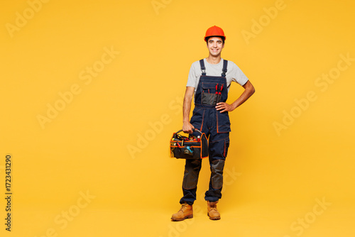 Full body young smiling happy employee laborer handyman man wear helmet overall hold bag with tools isolated on plain yellow background. Instruments for renovation apartment room. Repair home concept.