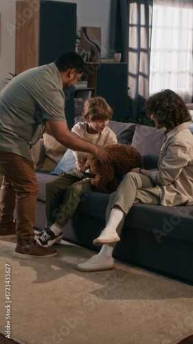 Vertical view of father bringing brown puppy home showing it to impressed son who petting dog with his mother while sitting on couch