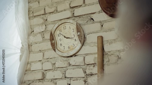 Old warped analog clock with distorted face hangs on white brick wall, ticking steadily as time passes, vintage style evokes surreal feeling of time slipping in rustic damaged indoor setting
