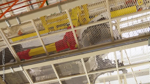 The undercarriage of a car body moves along an assembly line conveyor in an automobile factory. A low angle view from beneath the industrial conveyor belt with a protective metal grate.