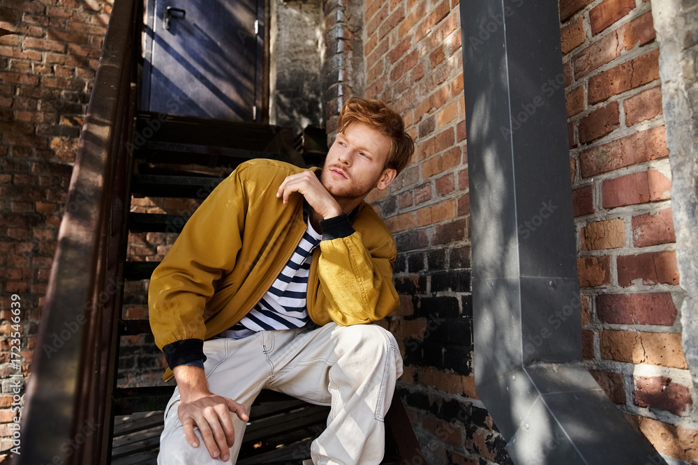 Fototapeta premium Handsome redhead man in stylish autumn outfit poses in city alleyway
