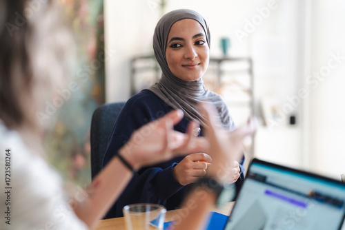 A young Muslim woman in a hijab smiles while listening intently during a meeting. She is engaged in a discussion with a colleague, with a laptop on the table.