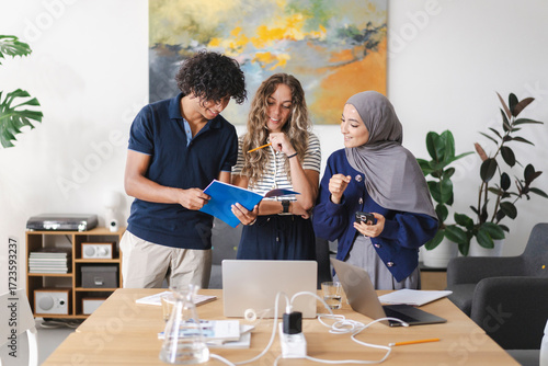 A diverse group of three young adults, including a man and two women, one wearing a hijab, collaborate around a table. They are smiling, discussing content in a blue notebook