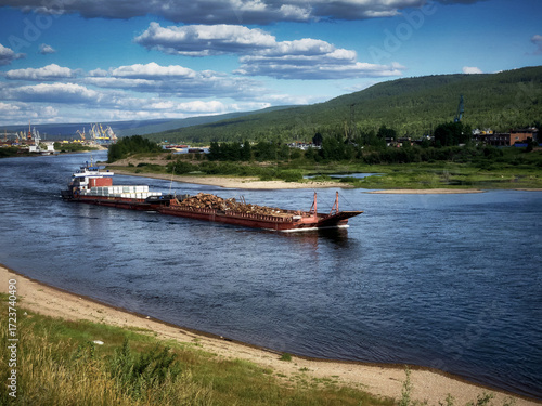 A warm summer day, a river, a self-propelled barge with various cargoes sailing along the river, cloudy sky