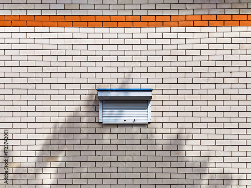 The wall is made of light brick, with a narrow strip of orange brick, in the middle of the wall there is a tiny window closed with blinds, a sketch from urban life
