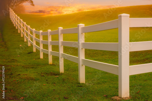  AI-Generated image of a white wooden fence stretching diagonally across a lush green field at sunset.
