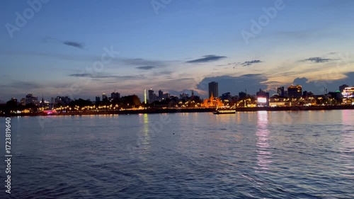 Sunset view of the Phnom Penh riverfront in Cambodia, featuring the illuminated Royal Palace boat sailing and surrounding skyline. Cultural heritage with modern high-rises blending concept
