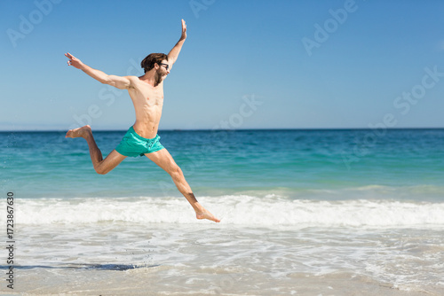 Man wearing turquoise swim shorts and dark sunglasses leaping above surf on sandy beach, copy space