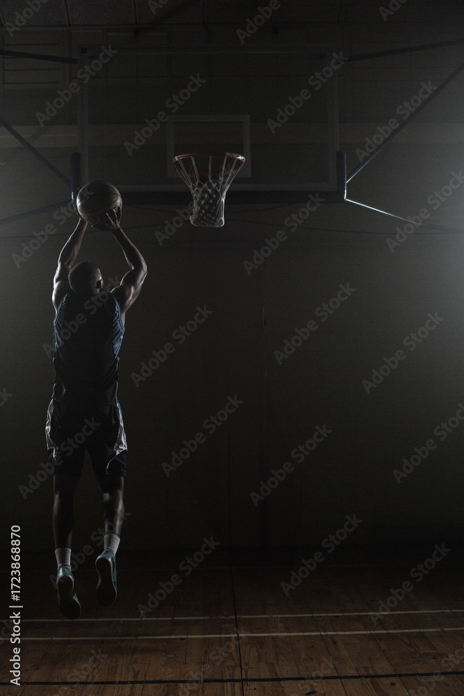 Fototapeta premium Basketball flying toward hoop in dimly lit gymnasium, spotlights casting dramatic shadows