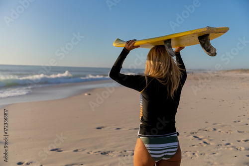 Female surfer carrying yellow surfboard overhead on sandy beach with footprints, copy space