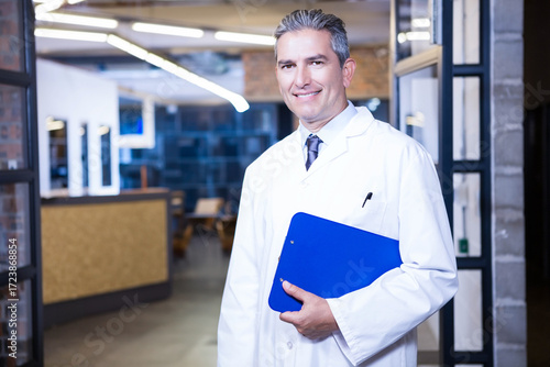 Professional wearing white lab coat holding blue clipboard standing in brick and glass corridor