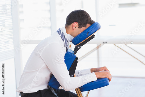 Man in business attire sitting on blue massage chair in wellness room leaning forward