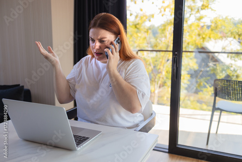 Angry woman sitting at desk with laptop, holding smartphone and gesturing in frustration. Concept of bad internet connection, online problems, stressful work, digital communication issues, remote job
