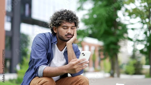 Lonely sad male student bored and using mobile phone while sitting on bench in campus space near university building. Tired young handsome man is depressed and in a bad mood, browses web on smartphone