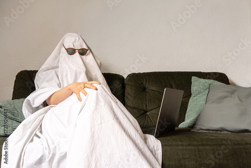 Person disguised as a ghost with a white sheet and sunglasses, lounging on a sofa with a computer on the side