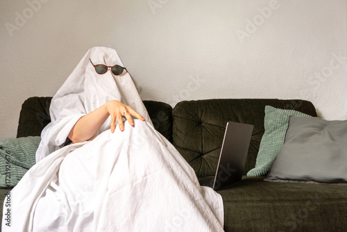 White sassy bed sheet ghost sitting on a couch with sunglasses and a laptop, concept of remote work with humor.