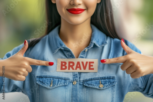 Young woman pointing to brave on her denim shirt, smiling confidently to express courage, self esteem and inner strength empowered, proud and ready to face the future