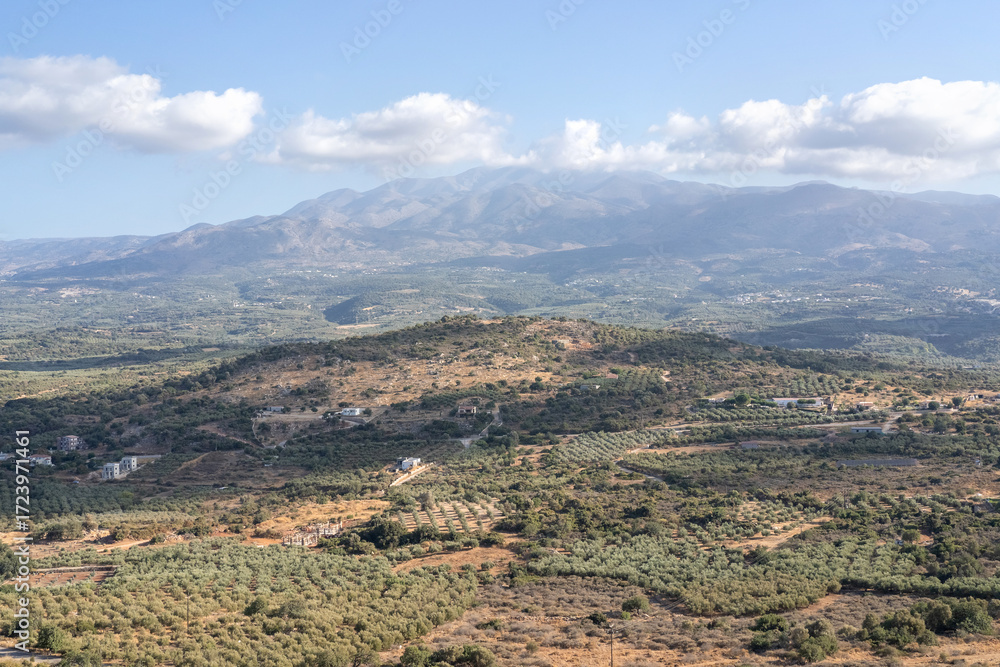 Fototapeta premium Beautiful panoramic view of the Cretan countryside with mountains, green valleys, olive groves, and a small traditional village under a bright blue sky with scattered clouds. Captured on a sunny day
