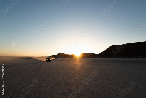 Two off-road vehicles drive through a dusty desert plain at sunset, leaving long shadows across the cracked earth, symbolizing adventure and freedom