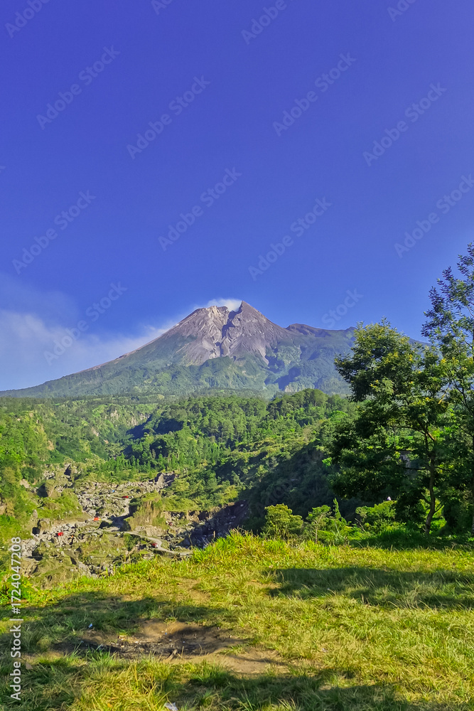 Fototapeta premium A close-up view of Mount Merapi from a distance with trees in the foreground. The panoramic beauty of Mount Merapi on a clear, midday day is clearly visible from a distance.