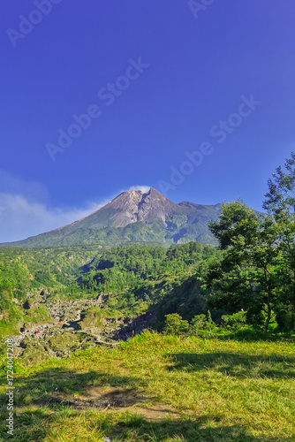 A close-up view of Mount Merapi from a distance with trees in the foreground. The panoramic beauty of Mount Merapi on a clear, midday day is clearly visible from a distance.