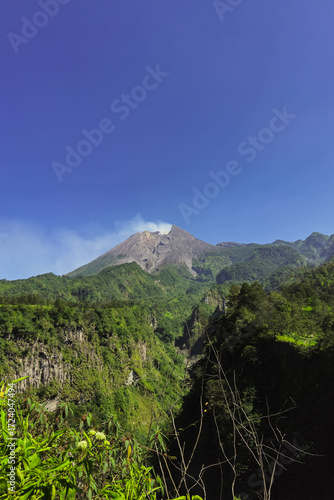 A close-up view of Mount Merapi from a distance with trees in the foreground. The panoramic beauty of Mount Merapi on a clear, midday day is clearly visible from a distance.