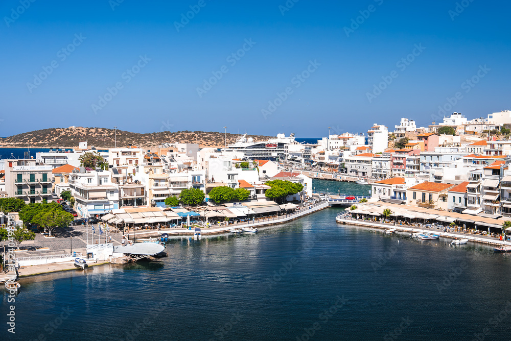 Obraz premium A serene waterfront town in Crete, Greece, featuring whitewashed buildings, terracotta rooftops, a small harbor with boats, and a curved pedestrian bridge.