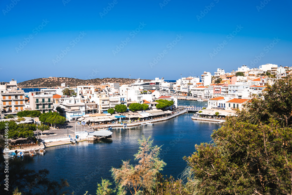 Obraz premium A serene waterfront town Agios Nikolaos in Crete, Greece, featuring whitewashed buildings, red tiled roofs, a small harbor with boats, and a backdrop of rolling hills.