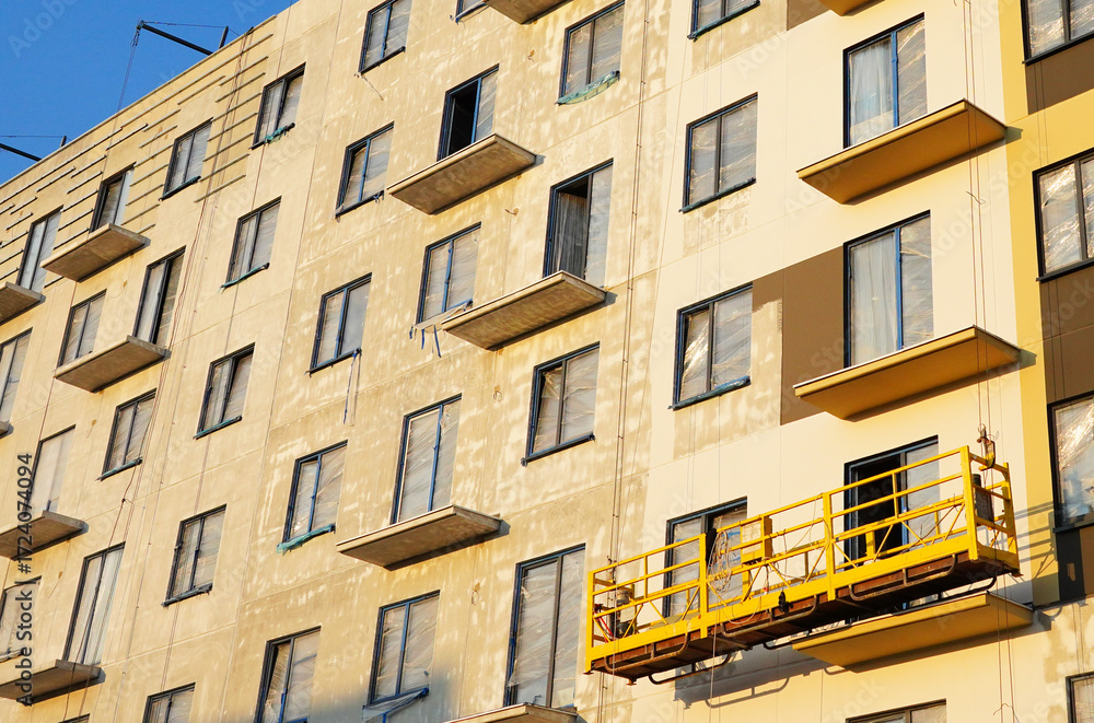 Fototapeta premium Suspended construction cradle on painting facade of building. Mobile scaffolding platform for construction work at height, facade cleaning, plastering. Insulation and wall painting, cladding.