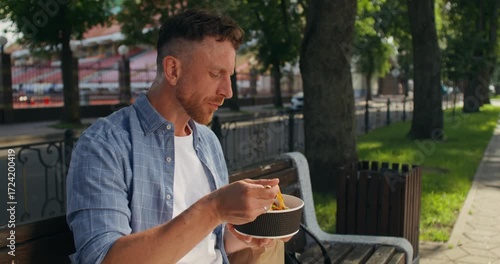 A man is eating takeout while sitting on a bench in the city. A man sits on a bench on a city street and takes out a takeaway from a paper bag.