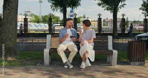 A man and a woman are talking sitting on a city bench. A man eats lunch with chopsticks, a woman drinks coffee.
