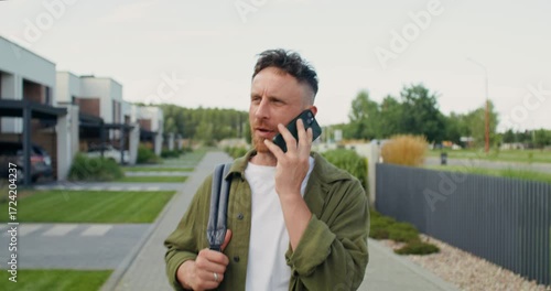 A man with a backpack on his shoulder is talking on a mobile phone while walking along a town street.