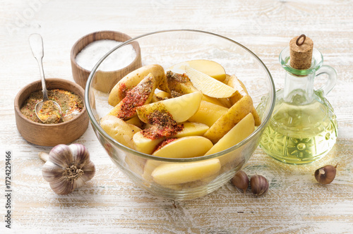 Fresh raw potato wedges in glass bowl with garlic, vegetable oil and colorful spices on rustic wooden table, cooking ingredients