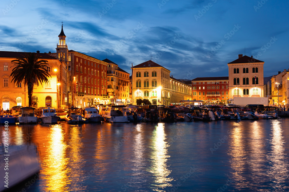 Naklejka premium Twilight and lantern light, largest port is located in Split. Private boats and yachts sway on waves, pedestrian tourists walk along embankment.