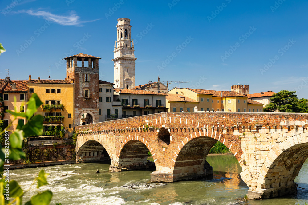 Obraz premium Scenic summer view of Ponte Pietra, oldest Roman bridge in Verona with graceful stone arches spanning Adige River, framed by houses draped in greenery and cathedral bell tower towering above skyline.