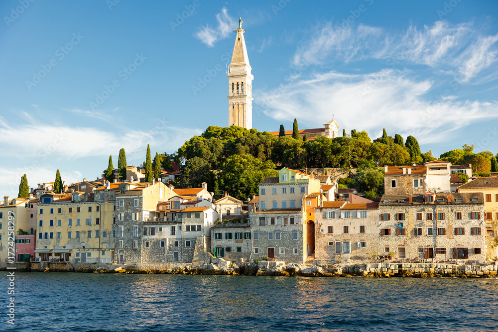 Fototapeta premium Bell tower of Church of St. Euphemia rises above city of Rovinj, citys dominant feature. Citys architecture and nature attract tourists. View of city from boat.