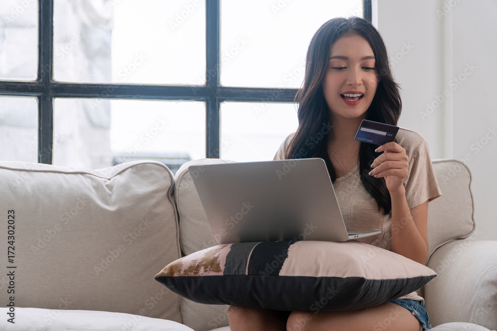 Naklejka premium Cheerful beautiful asian woman sitting on sofa with pillow using laptop browsing web online shopping payment credit card at apartment. Lifestyle technology purchase payment finance relaxation at home.