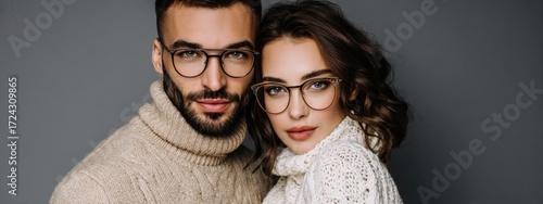 Elegant couple wearing glasses posing together against gray background. Man in beige turtleneck and woman in white knit top. Perfect for eyewear ads, optician campaigns, and professional branding.
