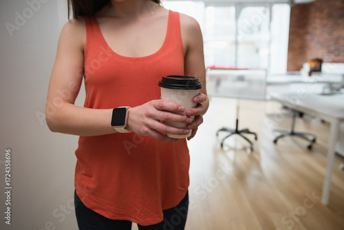 Asian worker wearing orange top and pants holding coffee cup in bright office with desks, chairs