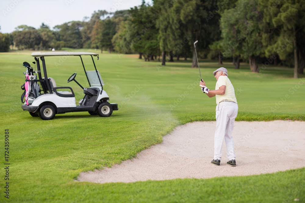 Obraz premium Senior man swinging club, hitting golf ball from bunker on golf course beside cart, copy space