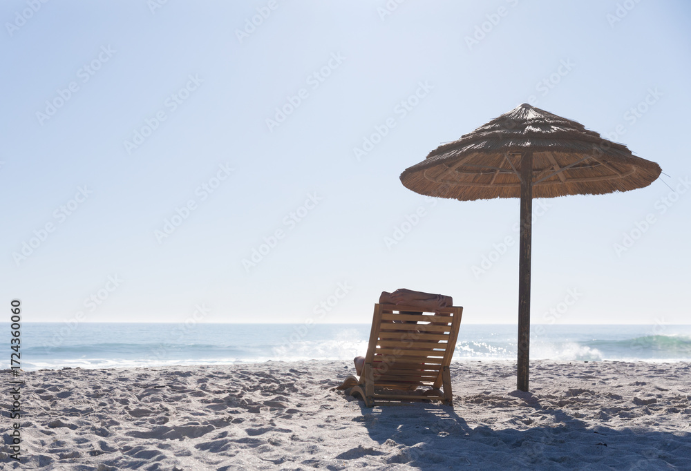 Fototapeta premium Wooden lounge chair draped red towel standing under straw parasol on beach facing sea, copy space